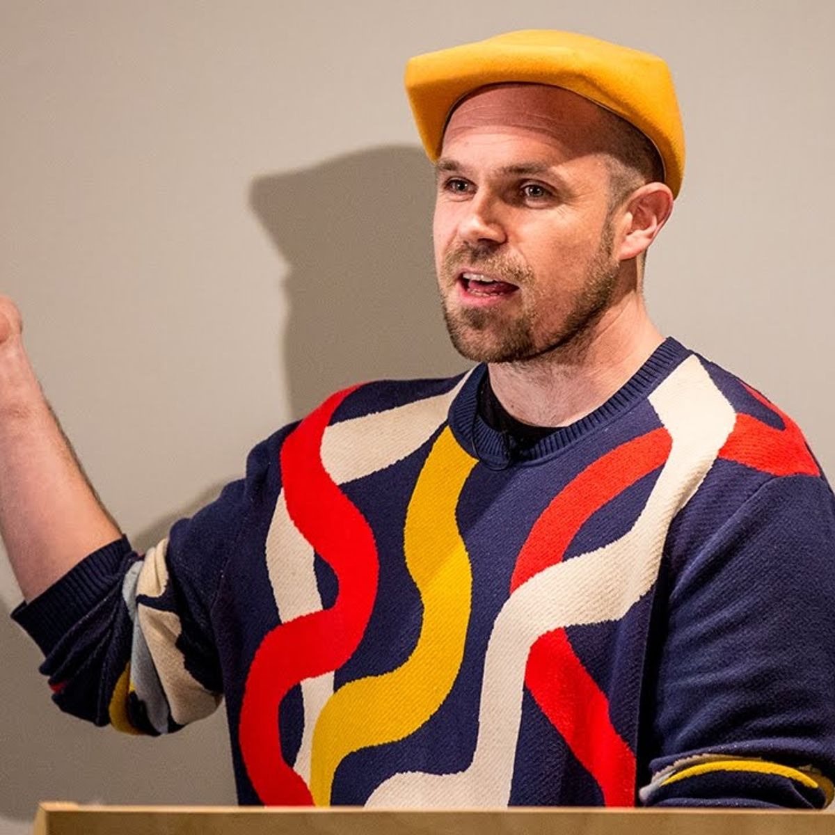 A man wearing a bright yellow beret and a colourful jumper with red, yellow, and white wavy lines gestures while speaking at a lectern—it’s clear his style says, “It Will Glow.”.