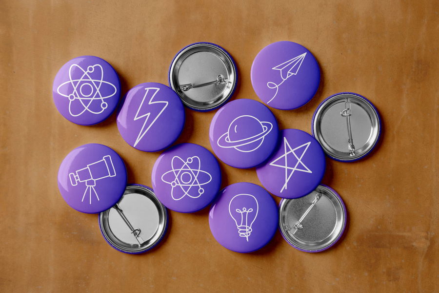 A group of purple pinback badges with white line drawings of science and space symbols, such as atoms, a telescope, a planet, a lightbulb, a lightning bolt, a paper aeroplane, and a star, on a wooden surface.