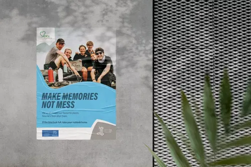 A poster on a grey wall shows five smiling people in outdoor gear cleaning up litter. The text reads, MAKE MEMORIES NOT MESS and encourages visitors to take their rubbish home from favourite places. A plant appears in the foreground.