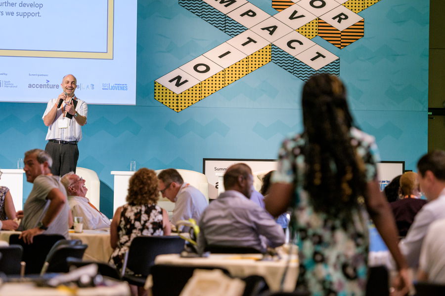 A man stands on stage speaking into a microphone at a conference, whilst audience members sit at round tables and one woman stands in the foreground facing him. Large text on the wall behind reads IMPACT 20-T.