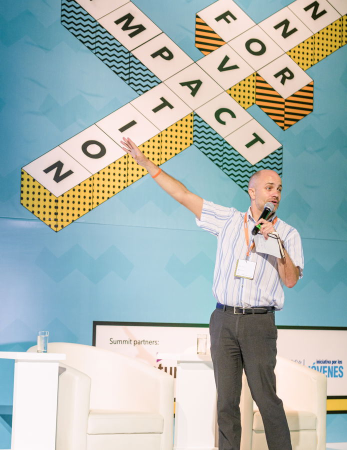 A man holding a microphone stands on stage, gesturing with one arm raised. Behind him, large geometric signs display the words IMPACT and CONTACT in a crossword puzzle style.