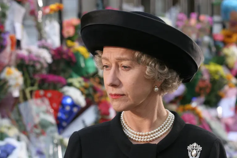 A woman dressed in black with a wide-brimmed hat, pearl necklace, and brooch stands sombrely before a wall of colourful flowers, evoking the elegance seen at The Queen’s Platinum Jubilee.