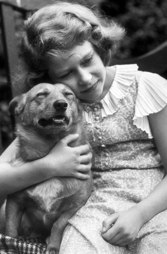 A young girl in a vintage dress hugs a small dog outdoors, her gentle smile reflecting the warmth of The Queen’s Platinum Jubilee celebrations, whilst the contented pup relaxes peacefully in her embrace.