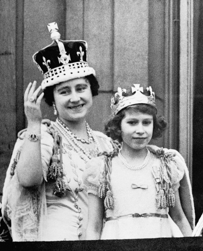 A woman and a young girl, dressed in ornate attire and sparkling jewellery, smile for the camera during The Queen's Platinum Jubilee. Both wear crowns; the woman waves gracefully whilst the girl stands proudly in front.