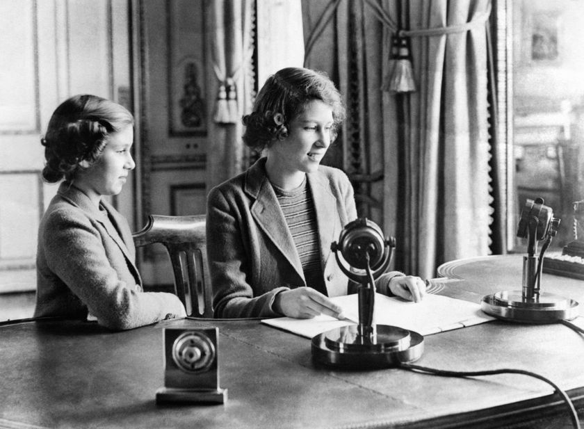 Two young girls sit at a desk with microphones and papers, dressed in formal jackets and appearing focused as they prepare to speak or read, with elegant curtains and décor in the background, celebrating The Queen’s Platinum Jubilee.