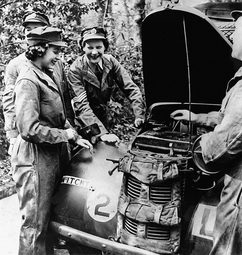 Four women in military uniforms smile and chat whilst examining an engine under an open vehicle bonnet, evoking the camaraderie of the Second World War era—a fitting tribute to service celebrated during The Queen's Platinum Jubilee.