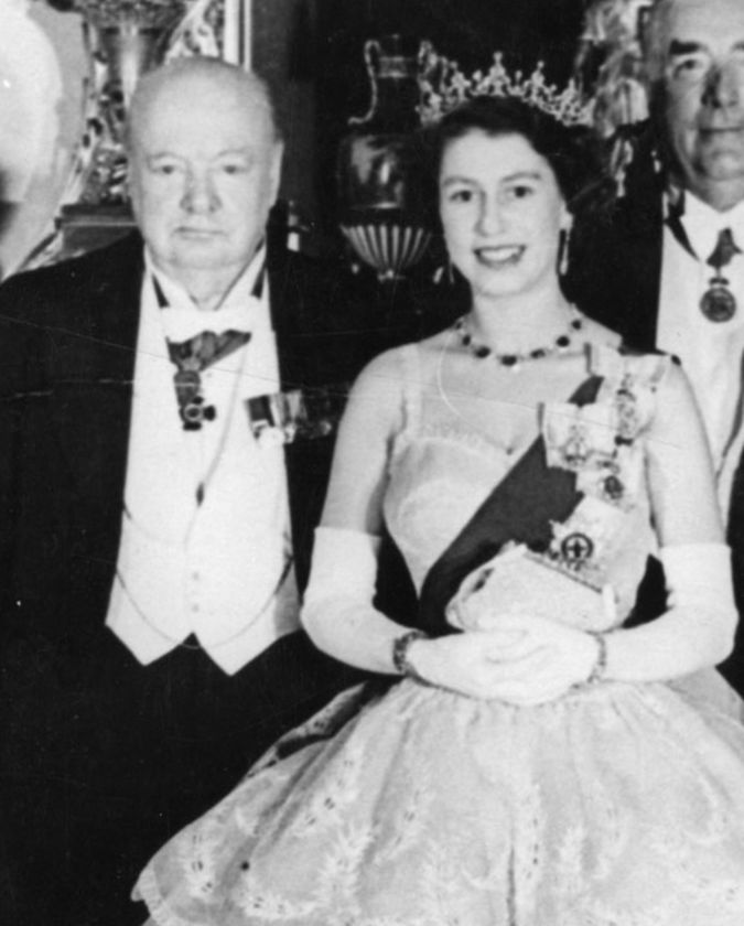 A black and white photo showing an older man in formal attire standing beside a young woman wearing a tiara, sash, and elegant gown, both smiling and dressed for a ceremonial event during The Queen’s Platinum Jubilee.