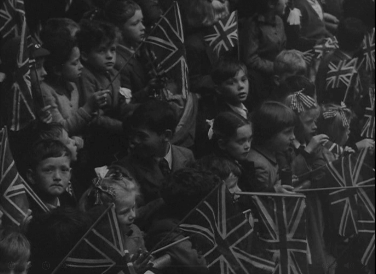 A large group of children sit closely together, many holding and waving Union Jack flags, likely celebrating The Queen's Platinum Jubilee. Most children are looking in the same direction, some with ribbons in their hair. The image is black and white.
