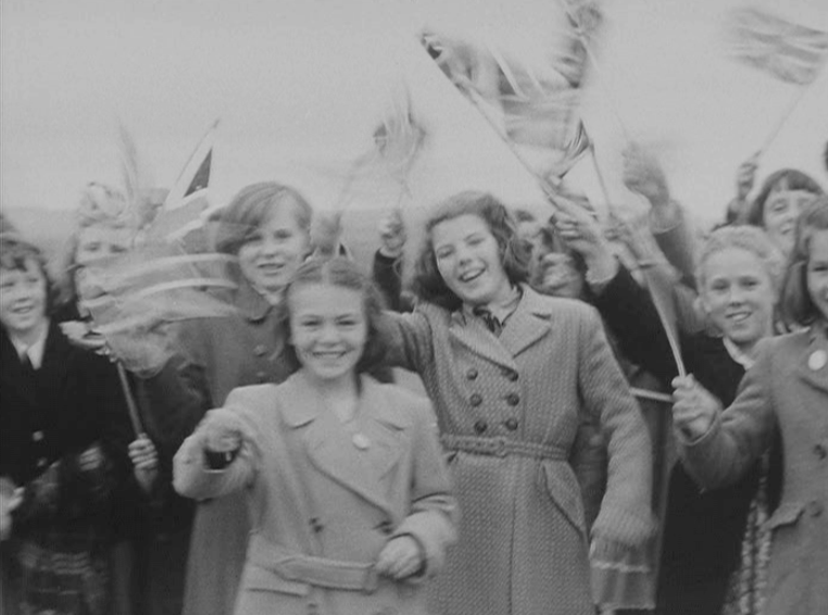 A group of smiling children wave flags and cheer, dressed in coats. The black and white photo captures their joyful celebration outdoors—perhaps marking a special event like The Queen’s Platinum Jubilee.