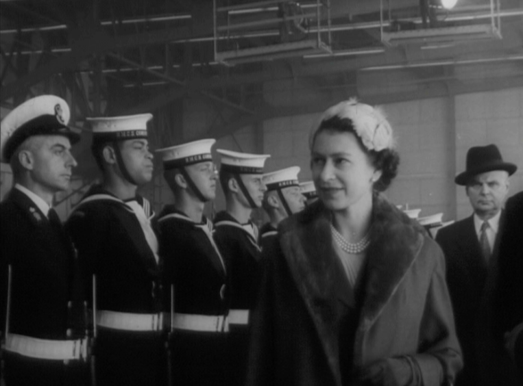 A woman in a fur coat and hat smiles whilst walking past a row of uniformed sailors standing at attention indoors, evoking scenes reminiscent of The Queen's Platinum Jubilee; several men in suits are visible in the background.