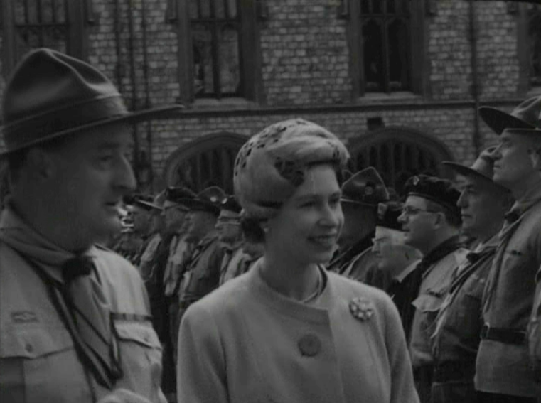 A woman in formal attire and a man in a scout uniform stand before uniformed men, with a stone building and arched windows behind them. The black-and-white image evokes the spirit of The Queen’s Platinum Jubilee.