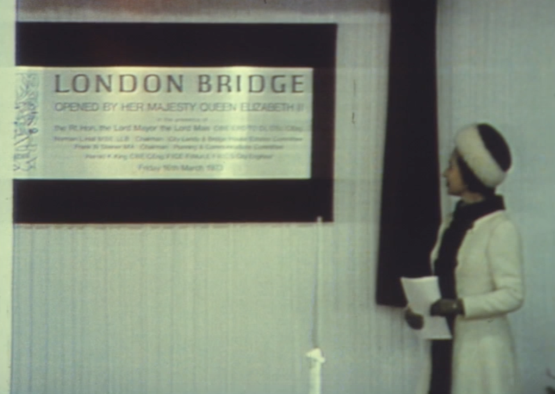 A woman in a white coat and hat stands next to a plaque that reads London Bridge, opened by Her Majesty Queen Elizabeth II, with additional dedication text—commemorating The Queen's Platinum Jubilee—mounted on a wall with a red curtain.