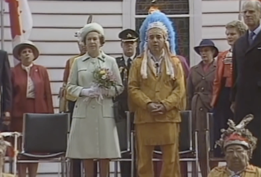 At The Queen's Platinum Jubilee outdoor ceremony, a woman in a light green coat and hat stands beside a man in a tan outfit with a blue feathered headdress, surrounded by officials and onlookers in both formal and traditional attire.