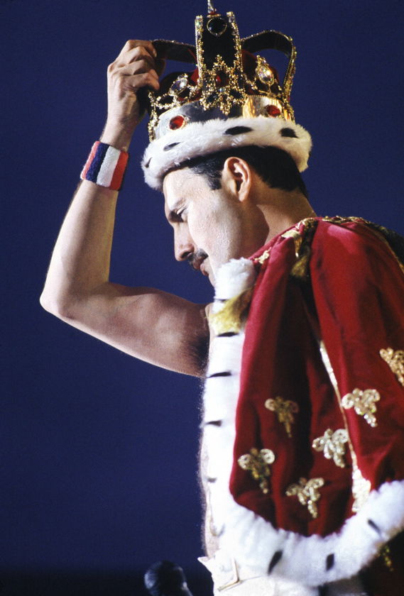 A performer wearing a jewelled crown and a red, gold-embroidered royal cloak holds the crown above his head on stage, set against a dark blue background, evoking the grandeur of The Queen’s Platinum Jubilee.