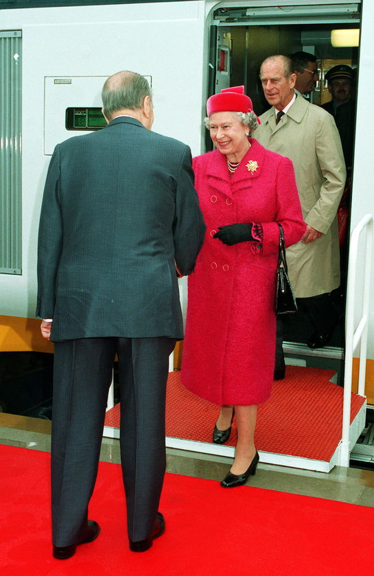 Queen Elizabeth II, dressed in a bright pink coat and hat, exits a train and shakes hands with a man in a suit on a red carpet, whilst Prince Philip stands behind her, during The Queen’s Platinum Jubilee celebrations.