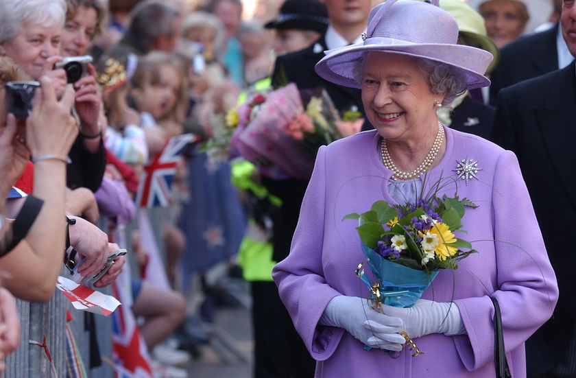 An elderly woman in a lavender coat and hat, holding a bouquet of flowers, smiles whilst walking past a crowd of people taking photos and waving flags during The Queen's Platinum Jubilee.