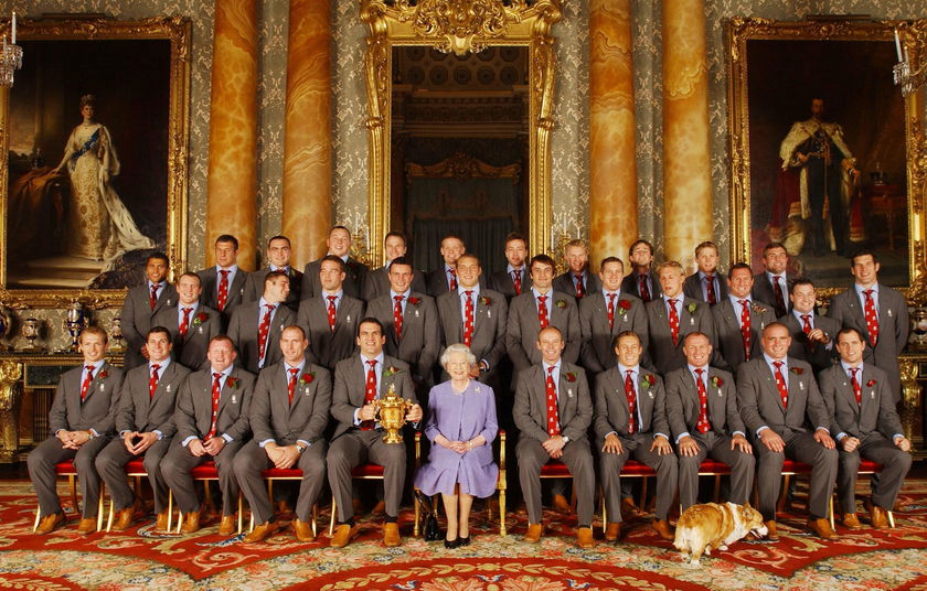 A group of men in grey suits with red ties pose for a formal photo in an ornate, gold-accented room. An elderly woman in lavender, celebrating The Queen's Platinum Jubilee, sits at the centre with a trophy and a dog at her feet.
