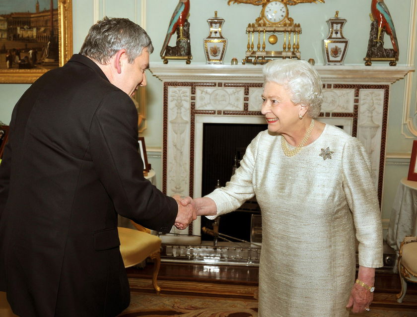 A man in a dark suit bows slightly whilst shaking hands with an older woman in a light-coloured dress in an ornate room, decorated for The Queen's Platinum Jubilee, with a fireplace, clocks, and elegant items in the background.