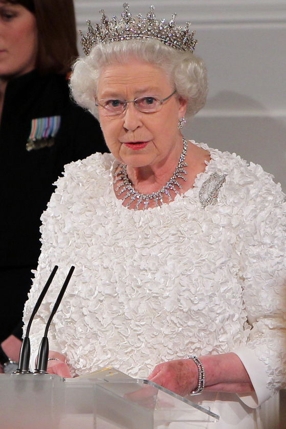 An older woman, dressed in a white textured dress with a crown and glasses, stands at a lectern for The Queen's Platinum Jubilee. Adorned with a diamond necklace and brooch, she addresses the audience as microphones capture her words.
