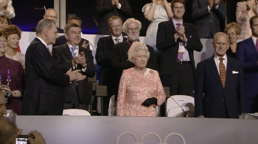 Queen Elizabeth II stands in the centre wearing a pink dress as people around her applaud during an Olympic event, with the Olympic rings in the foreground—a fitting tribute ahead of The Queen's Platinum Jubilee.