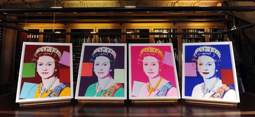 Four colourful pop art portraits of Queen Elizabeth II, each with vibrant colour schemes, are displayed side by side on easels in front of bookshelves, celebrating The Queen's Platinum Jubilee in a lively library setting.