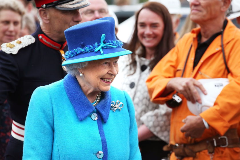 An elderly woman in a bright blue coat and matching hat smiles among a group of people, including a person in an orange boilersuit holding a white hard hat, as they celebrate the Queen's Platinum Jubilee in a cheerful and friendly atmosphere.