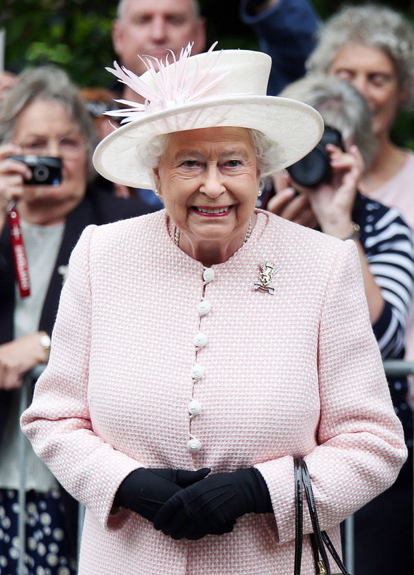 An elderly woman in a pale pink outfit, white hat with feathers, and black gloves smiles whilst standing outdoors during The Queen's Platinum Jubilee, surrounded by a small crowd, some of whom are taking photos.