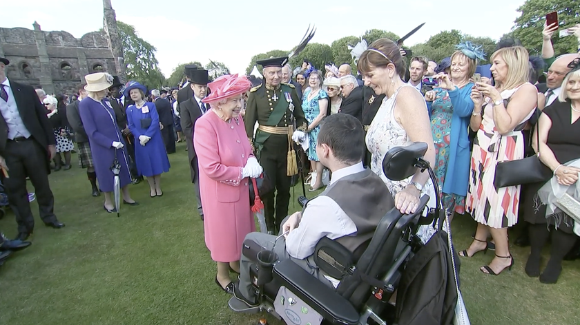 A woman in a bright pink coat and hat greets a man in a wheelchair on a lawn during The Queen’s Platinum Jubilee, surrounded by people in formal attire, with a historic stone building and trees in the background.