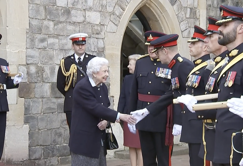 An elderly woman in a dark coat shakes hands with a uniformed officer outside a stone building, as several others in military uniforms stand nearby, saluting and observing during The Queen's Platinum Jubilee.