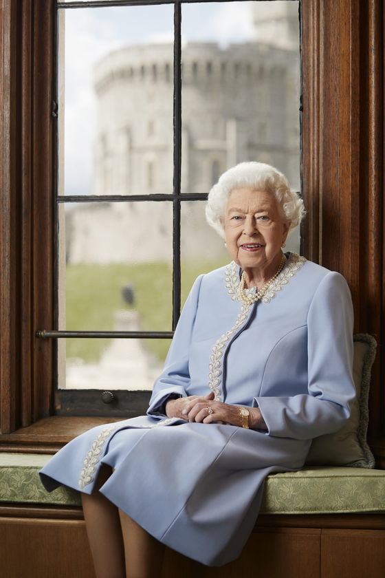 An elderly woman with white hair, dressed in a light blue dress with white embroidery, sits on a cushioned bench by a window. Outside, a stone castle stands proudly—an elegant nod to The Queen’s Platinum Jubilee.