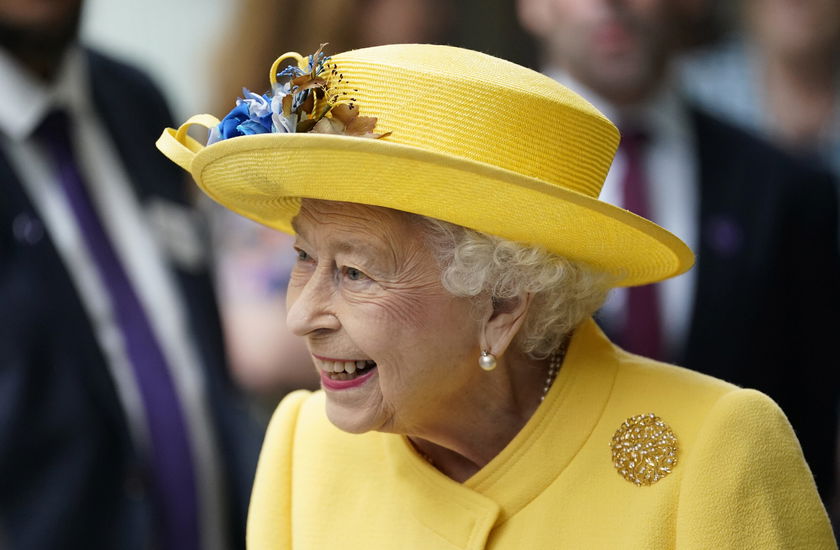 An elderly woman wearing a bright yellow hat with blue and yellow flowers and a matching yellow coat smiles whilst looking to the side during The Queen's Platinum Jubilee. People and blurred figures are visible in the background.