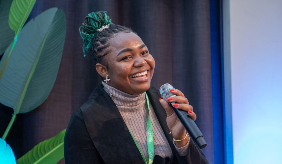 A woman with braided green hair, wearing a grey polo neck and a black blazer, smiles whilst holding a microphone. She has a green lanyard round her neck and is sitting in front of a plant and a dark curtain.