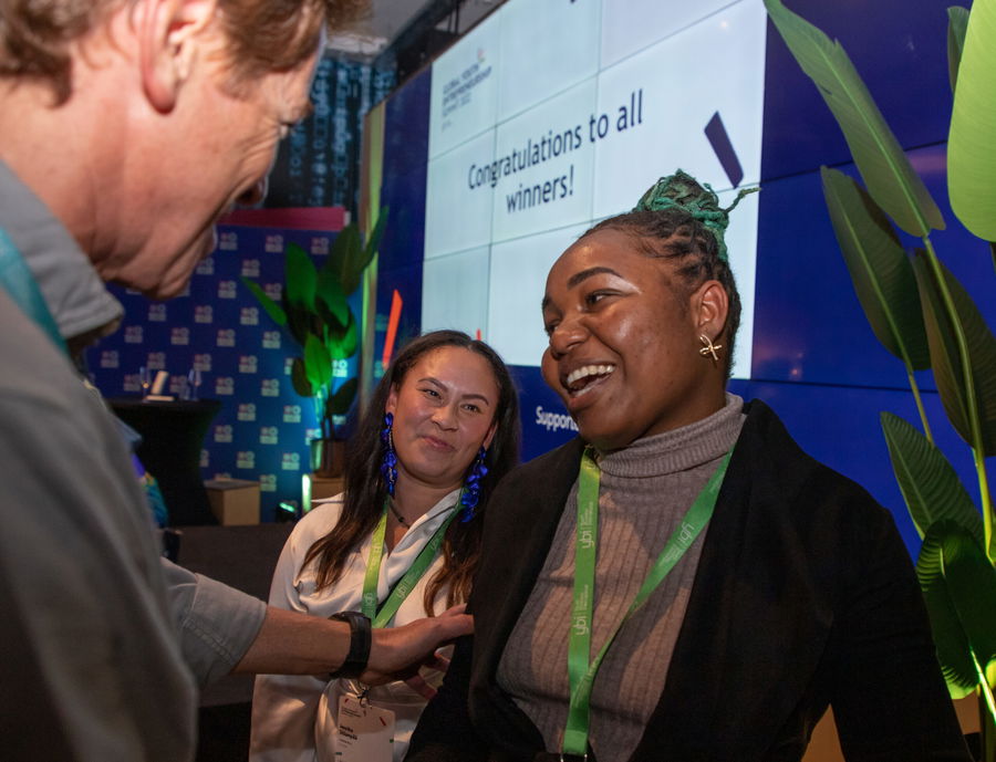 Three people interact at an event; two women smile, one wearing a green lanyard and plaits, while a man gestures. A screen in the background reads Congratulations to all winners!.