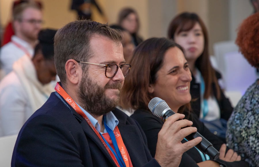 A man with glasses holds a microphone and smiles whilst seated next to a woman, also smiling, at an event. Other attendees are visible in the background.