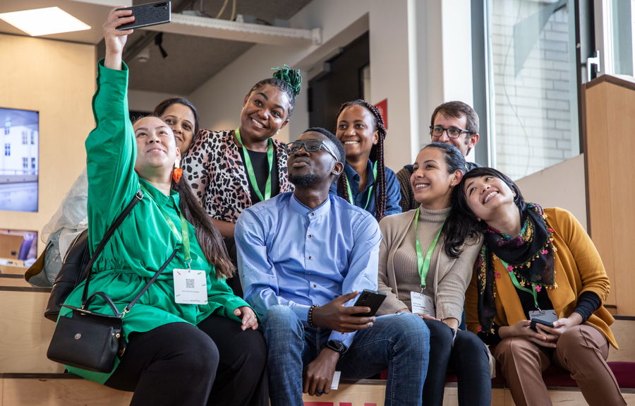 A diverse group of eight people sit closely together indoors, smiling and posing for a selfie. Most wear conference badges and green lanyards. Natural light comes through a window in the background.