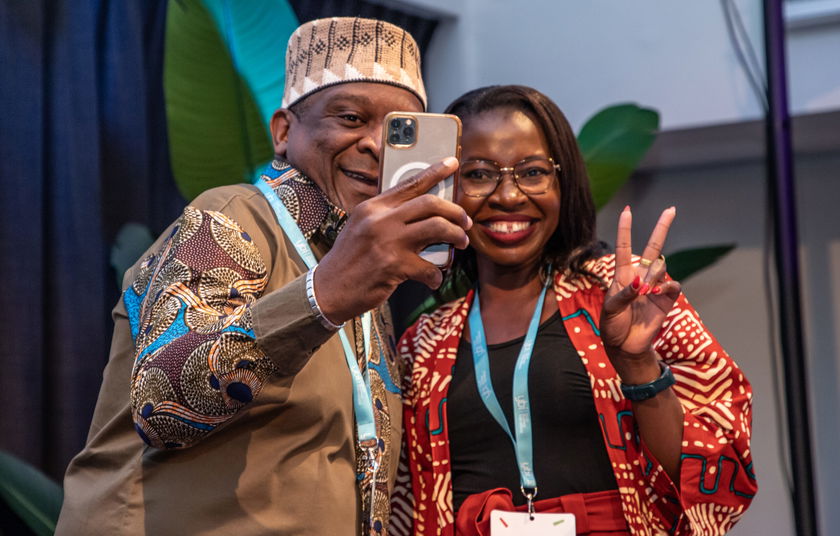 Two people smiling and posing for a selfie; the man holds a mobile while the woman makes a peace sign. Both wear colourful, patterned clothing and conference lanyards. Indoor setting with large green leaves in the background.