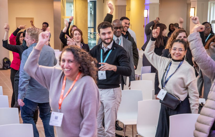 A group of people in a bright conference room stand and smile whilst raising their arms, participating in an interactive activity. Some wear conference badges and appear engaged and cheerful.