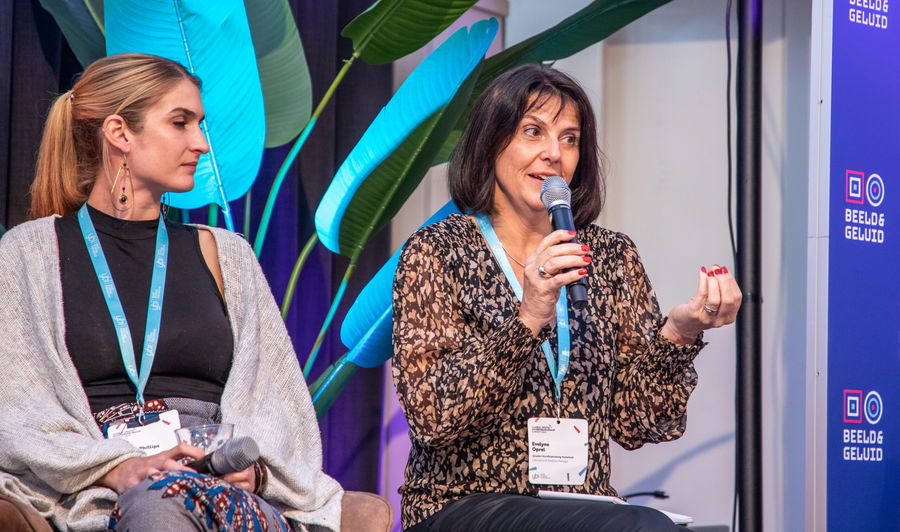 Two women sit on a panel; the woman on the right speaks into a microphone while gesturing, and the woman on the left listens. Both wear name badges and blue lanyards, with plant leaves visible in the background.