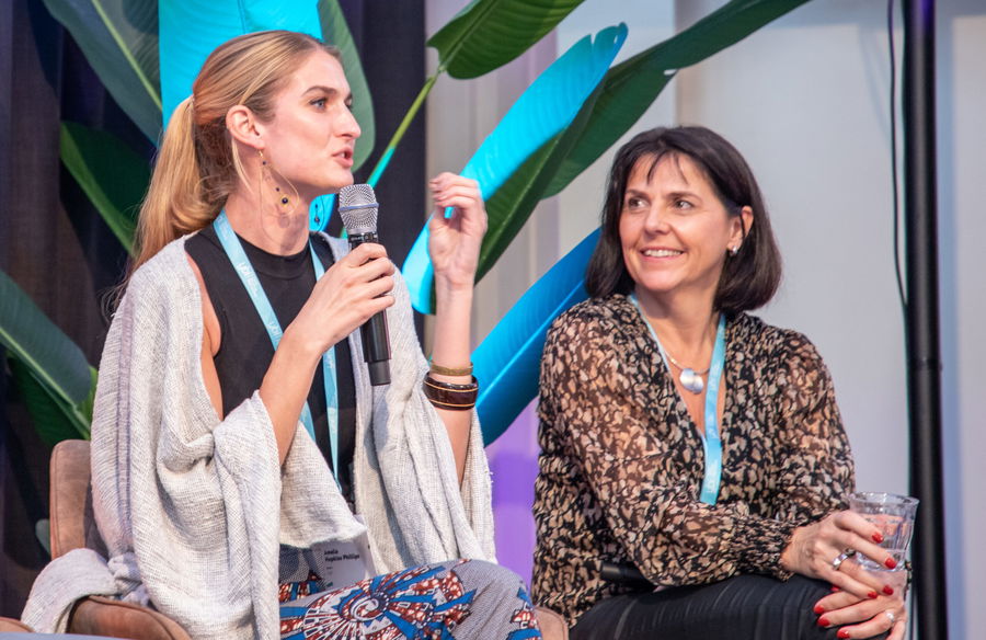 Two women sit on a panel. One, holding a microphone, speaks animatedly whilst the other smiles and listens. There are large green plants and blue lighting in the background.
