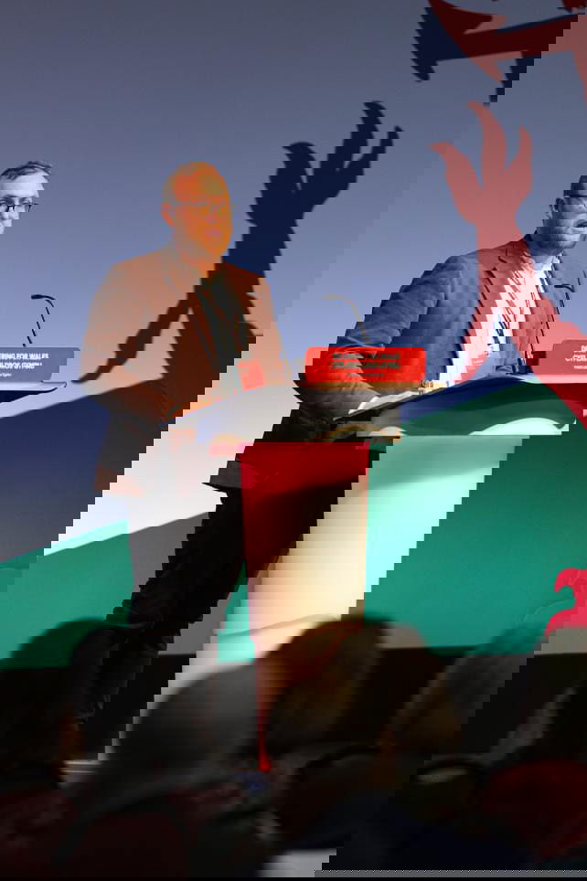 Jamie Green, in a suit jacket, stands at a lectern speaking on stage at a conference. The lectern displays a red sign reading Delivering For Wales, with a stylised dragon in the background and audience members seated in front.