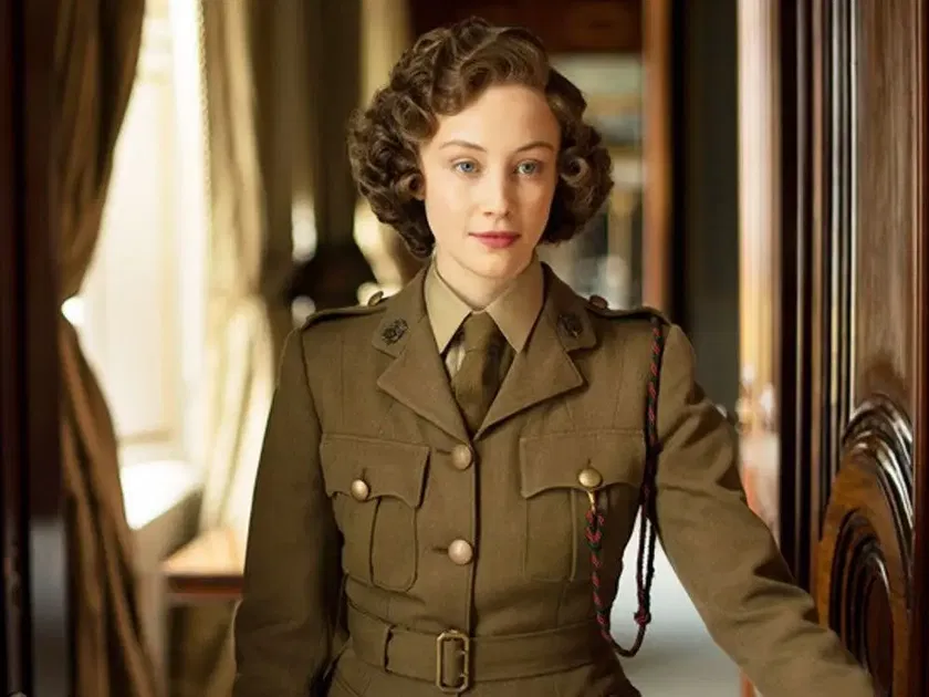 A woman with short curly hair wears a vintage military uniform with buttons and insignia, standing indoors beside a wooden doorway as sunlight streams through curtains—a look that echoes the timeless elegance of The Queen’s Platinum Jubilee.