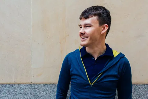 A smiling man with short brown hair, wearing a blue zip-up hoodie over a navy shirt, stands against a light beige and grey textured wall, looking to the right.