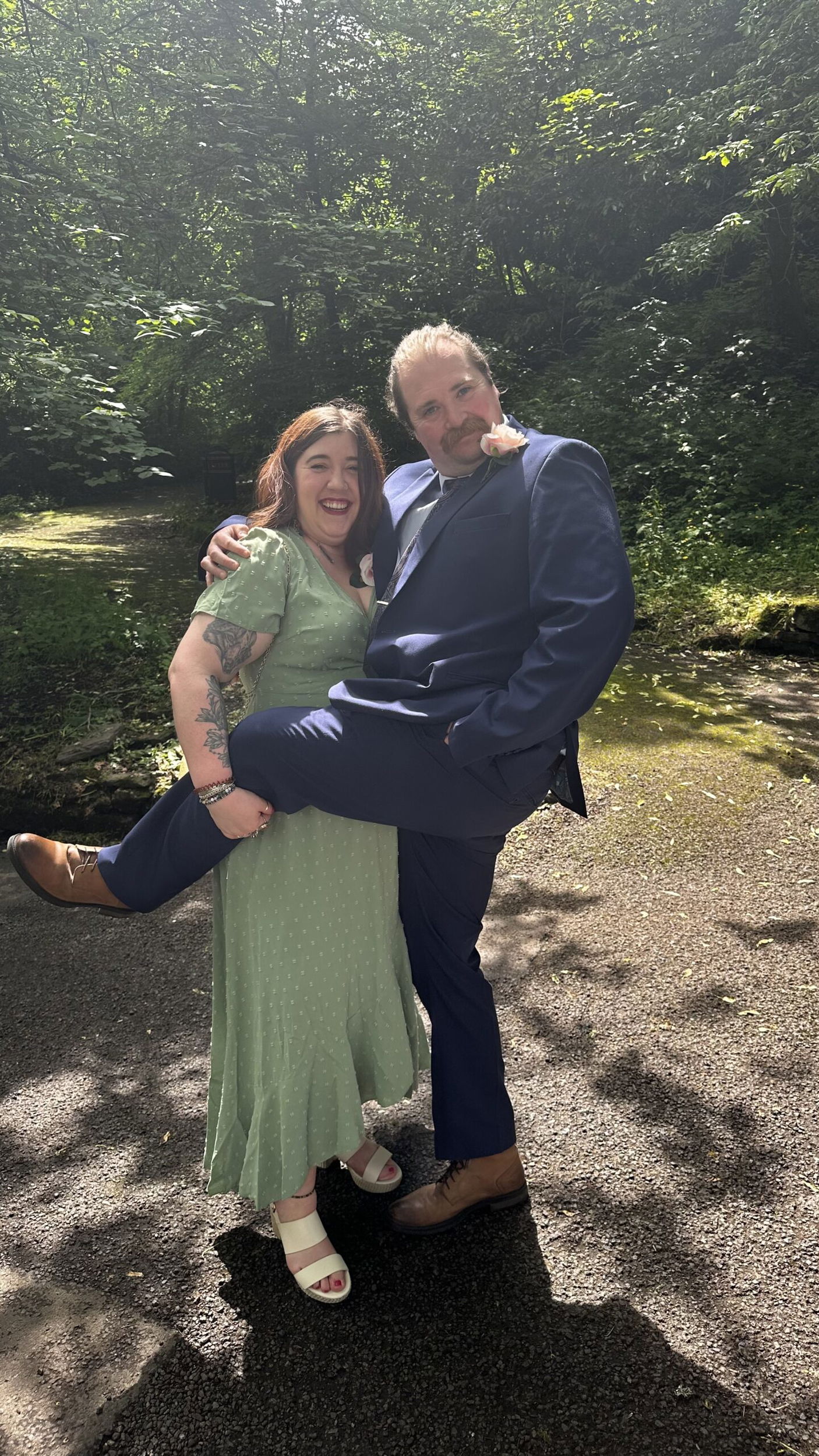 A woman in a light green dress and a man in a blue suit pose playfully outdoors; she smiles whilst holding his raised leg, and sunlight filters through the trees behind them.