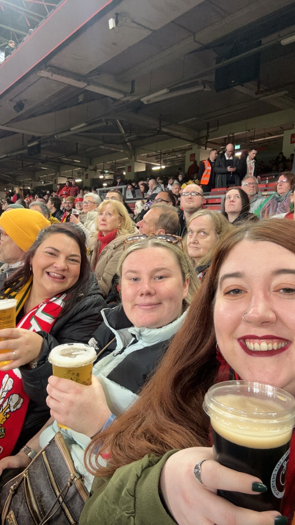 Three women smiling and holding cups of beer at a stadium, surrounded by other spectators. The atmosphere is lively, and they appear to be enjoying a sporting event.