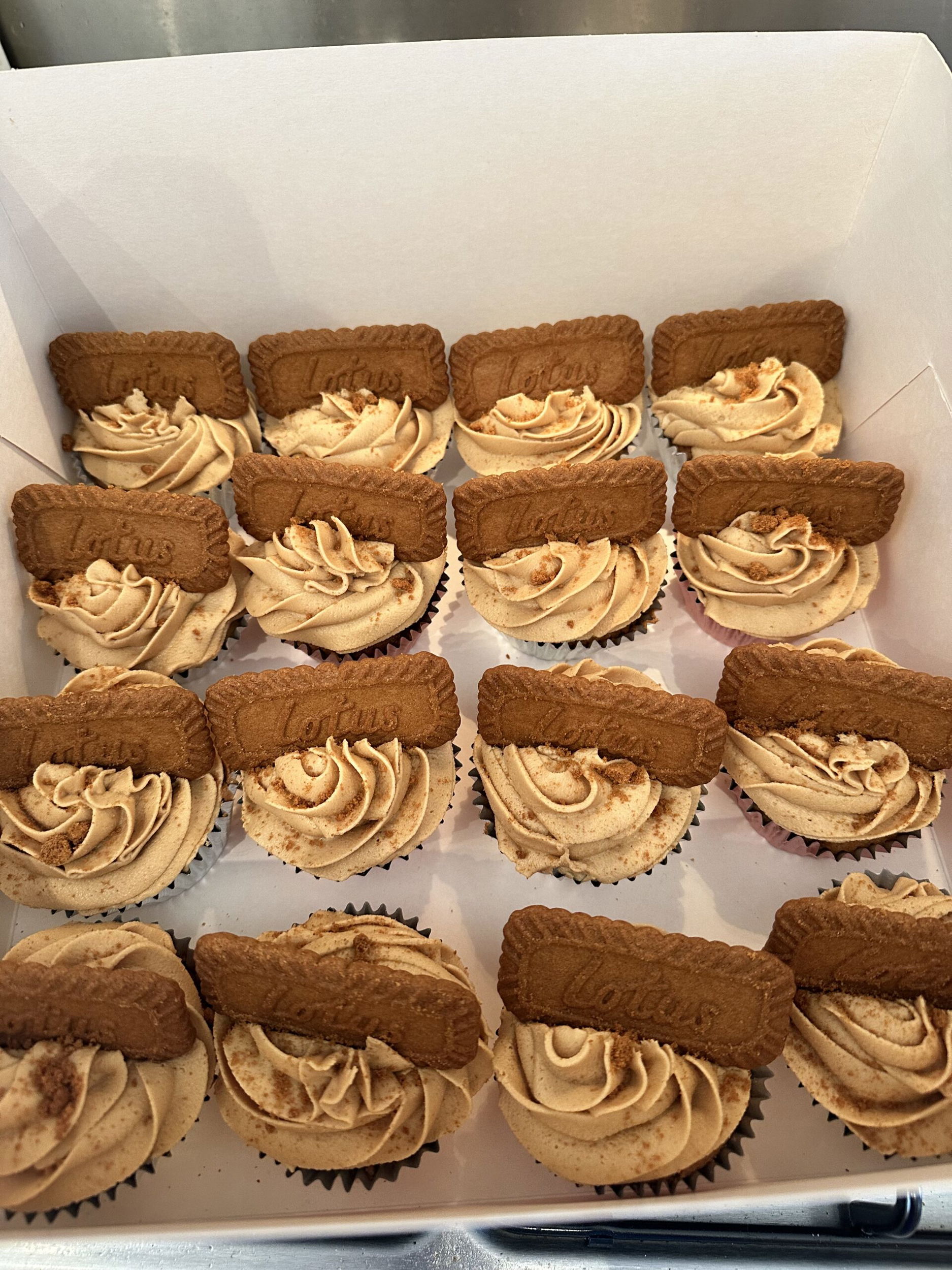 A box with rows of cupcakes topped with swirled light brown icing and a Lotus Biscoff biscuit on each one.