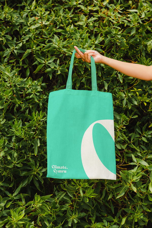 A hand holds a green and white tote bag with the words Climate. Cymru printed on it, against a lush background of green leafy plants.