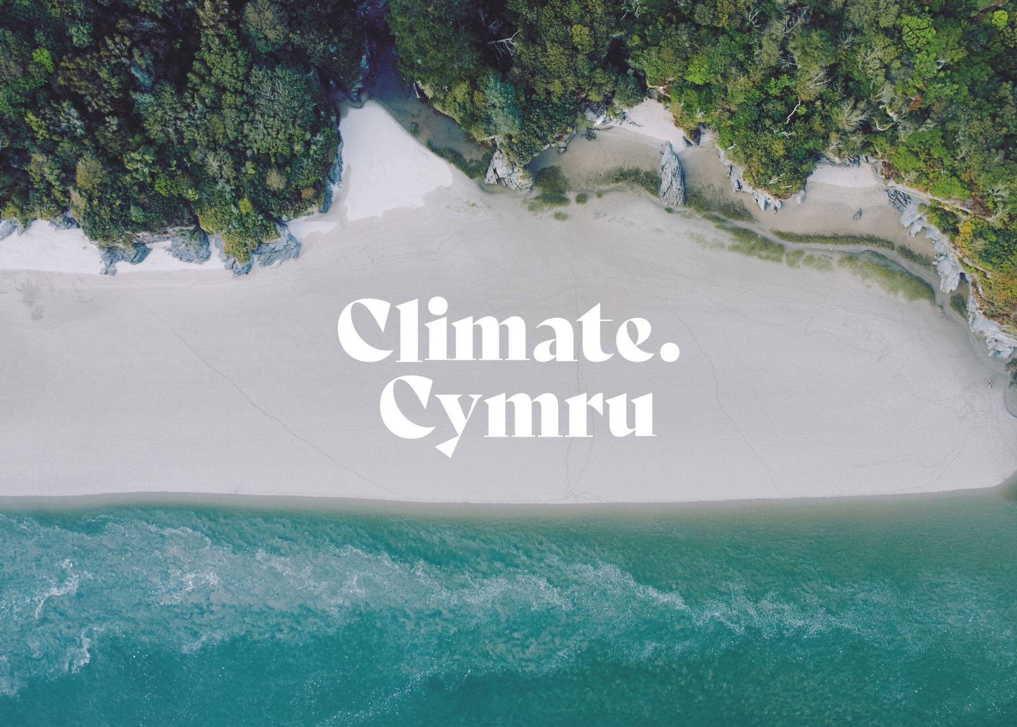 Aerial view of a sandy beach bordered by green trees and blue water, with the words Climate. Cymru in bold white text across the centre.