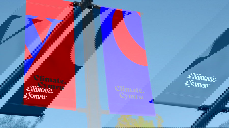 Two vertical banners on a pole read Climate. Cymru in white text; one banner is red with a blue design, the other blue with a red design. The sky is clear and blue in the background.