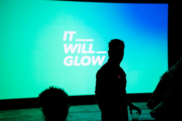 A person in silhouette stands before a large blue-green screen displaying the words IT WILL GLOW in white text at the Cardiff Design Festival. Other indistinct figures are visible in the foreground.