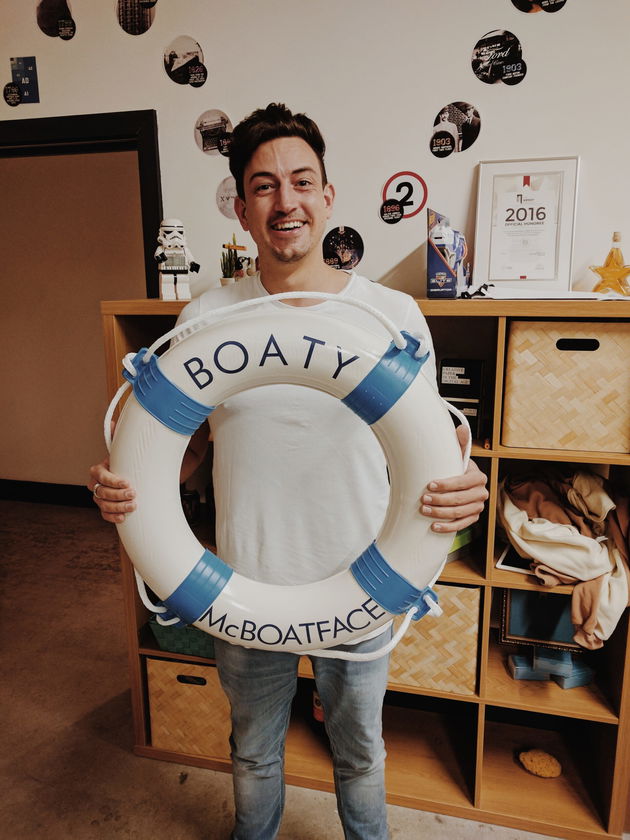 A smiling man stands indoors holding a white lifebuoy labelled Boaty McBoatface. Shelves with baskets, books, and wall decorations are in the background.